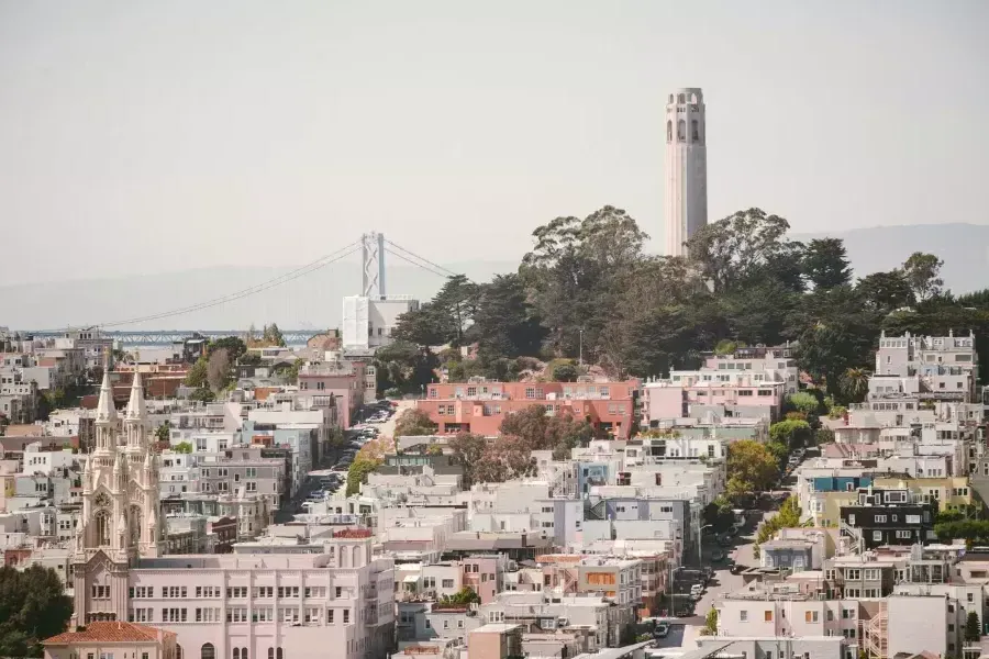 San Francisco's Coit Tower is pictured with the Bay Bridge in the background and a hill covered in houses in the foreground.